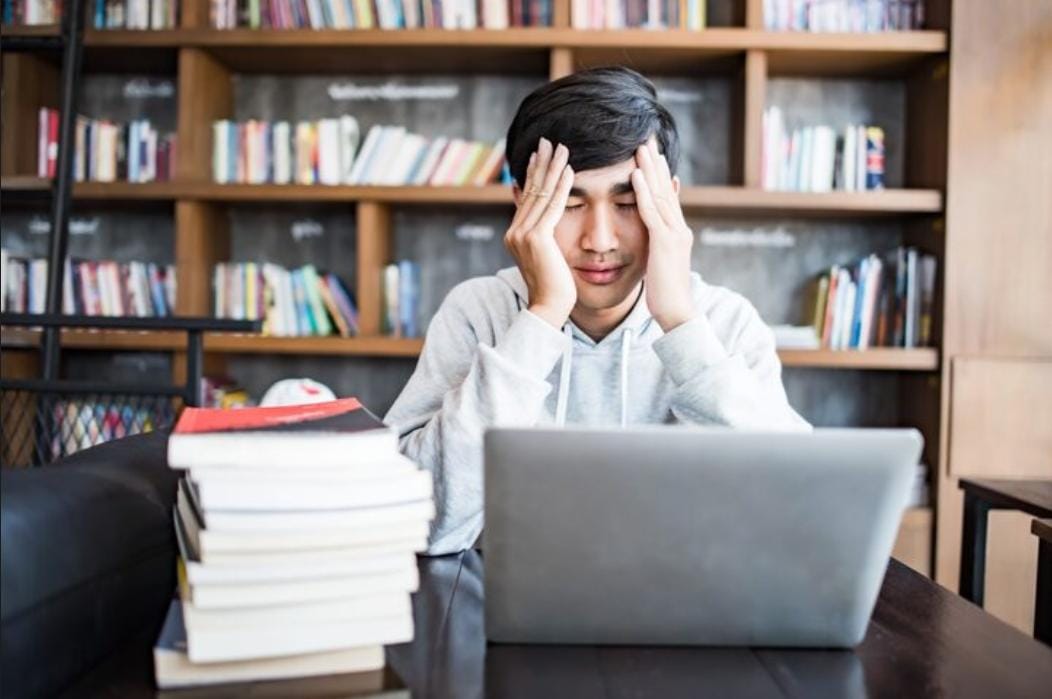 Mahasiswa belajar dengan tumpukan buku dan laptop
